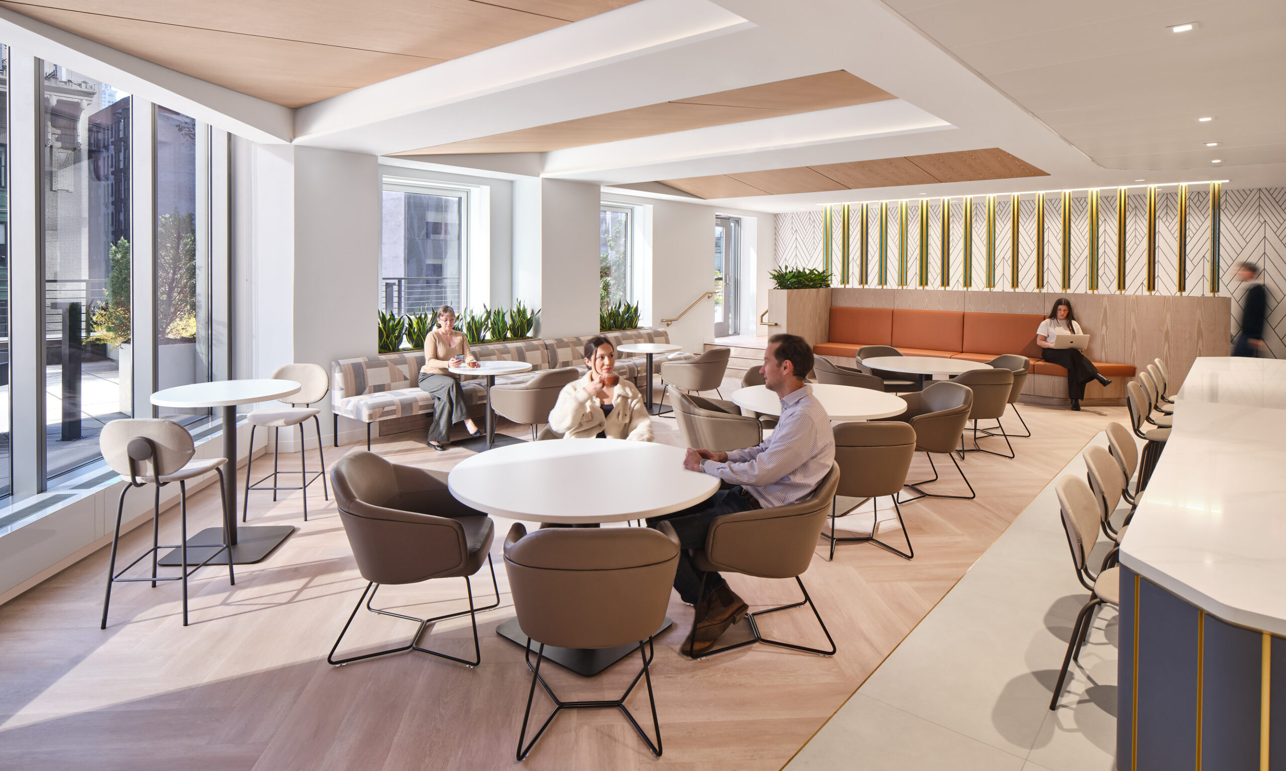 Bright and airy communal space featuring modern, circular tables surrounded by comfortable, padded chairs. Large windows allow natural light to flood the area, complemented by lush indoor plants. A cozy seating nook with a wooden wall panel adds warmth and texture, enhancing the inviting ambiance for collaboration and relaxation.