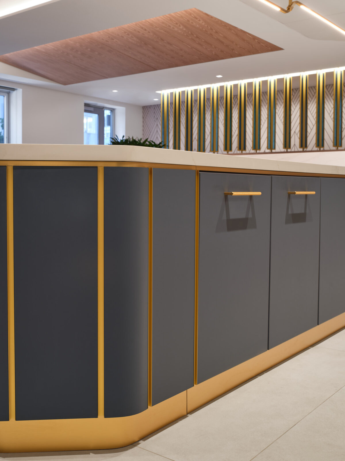 Sleek kitchen cabinetry features a modern design with dark gray finishes and striking gold accents. The curved edges of the cabinets create a dynamic flow, complemented by a sophisticated countertop. The back wall showcases an elegant geometric pattern illuminated by ambient lighting, enhancing the overall contemporary aesthetic.