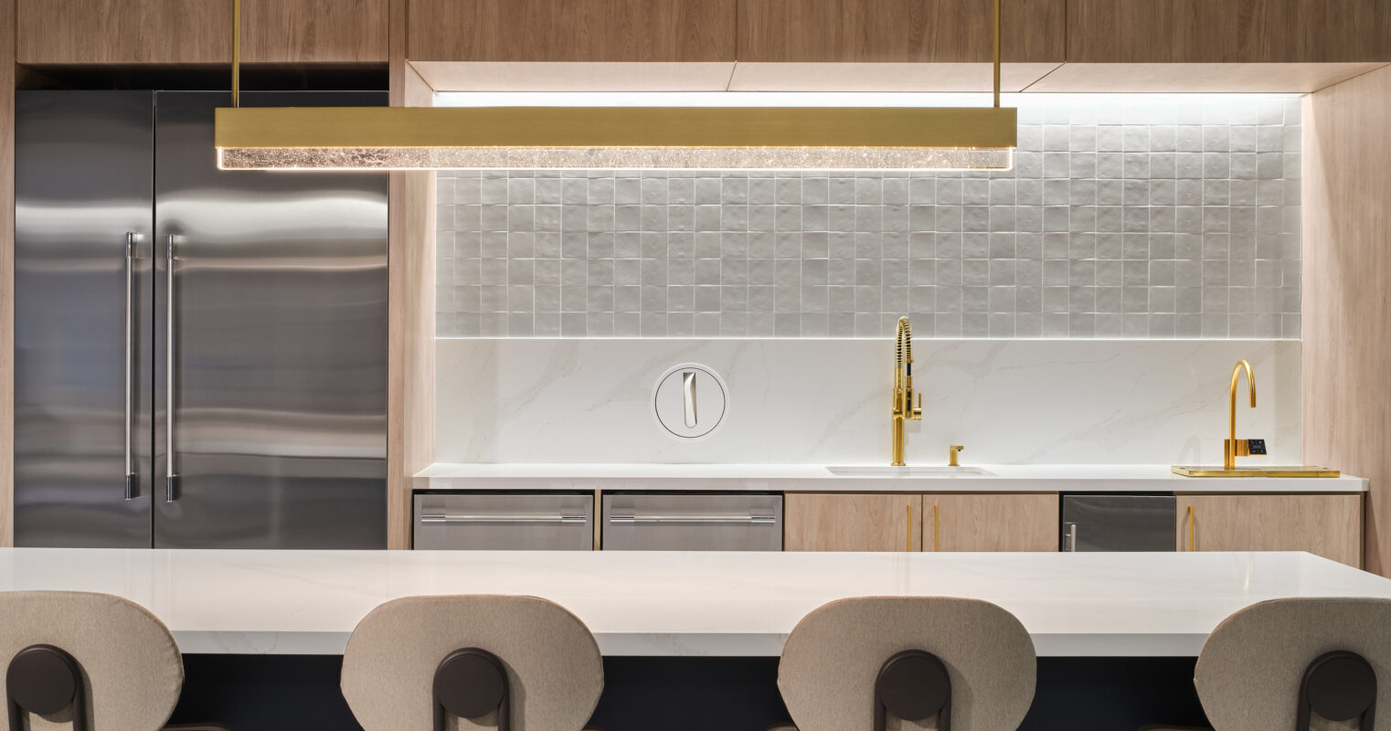 Modern kitchen featuring sleek wood cabinetry with a warm finish, complemented by stainless steel appliances. A striking white countertop with subtle veining contrasts with textured tile backsplash. Gold accents on the faucet and pendant lighting add a touch of elegance, while circular upholstered stools enhance comfort and style.