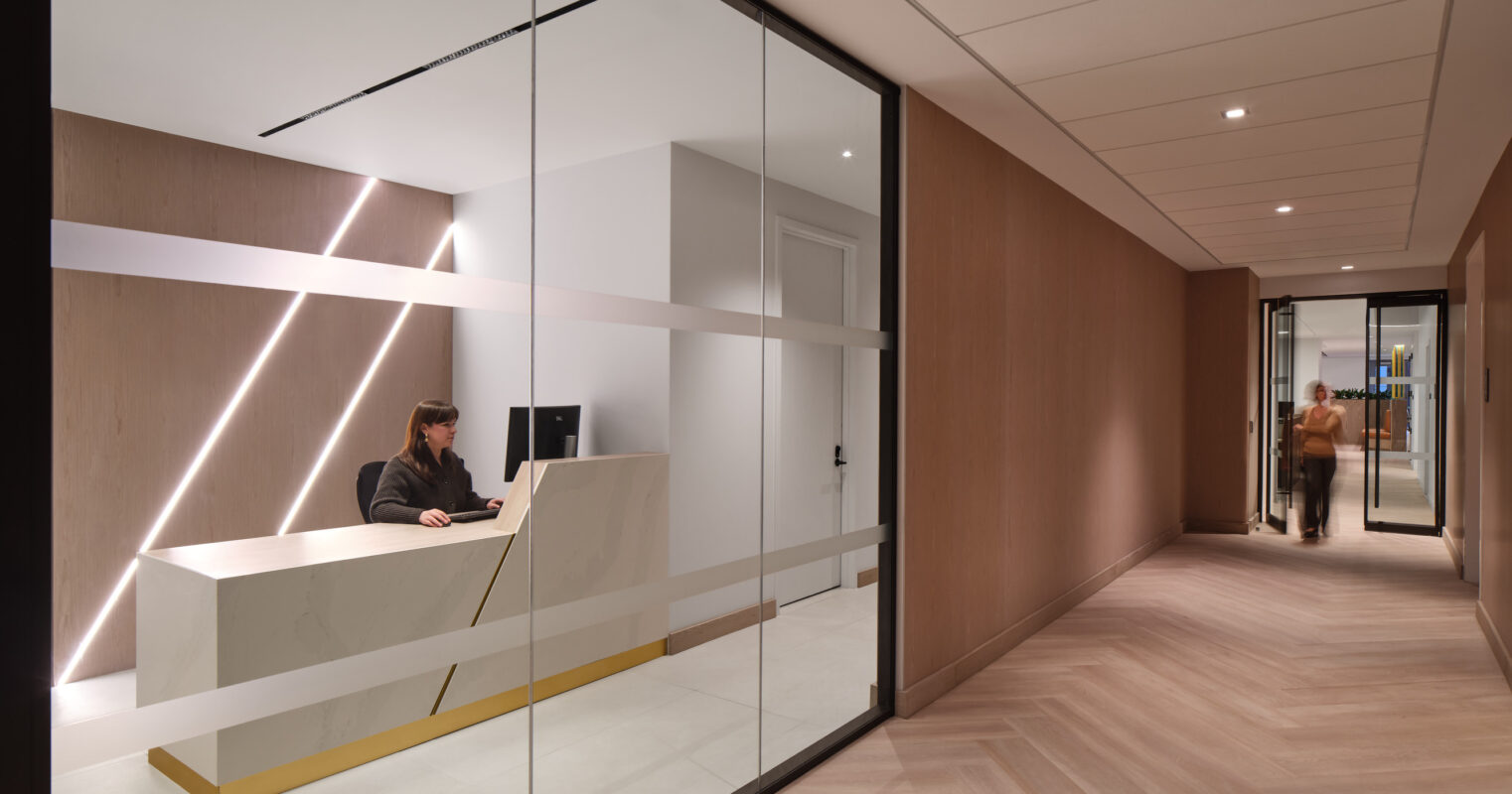 Sleek reception area featuring a minimalist desk with a striking white and gold design. Glass partitioning enhances openness while wooden paneling adds warmth. Soft, recessed lighting highlights the geometric lines on the wall, creating a welcoming atmosphere for visitors transitioning from the corridor.