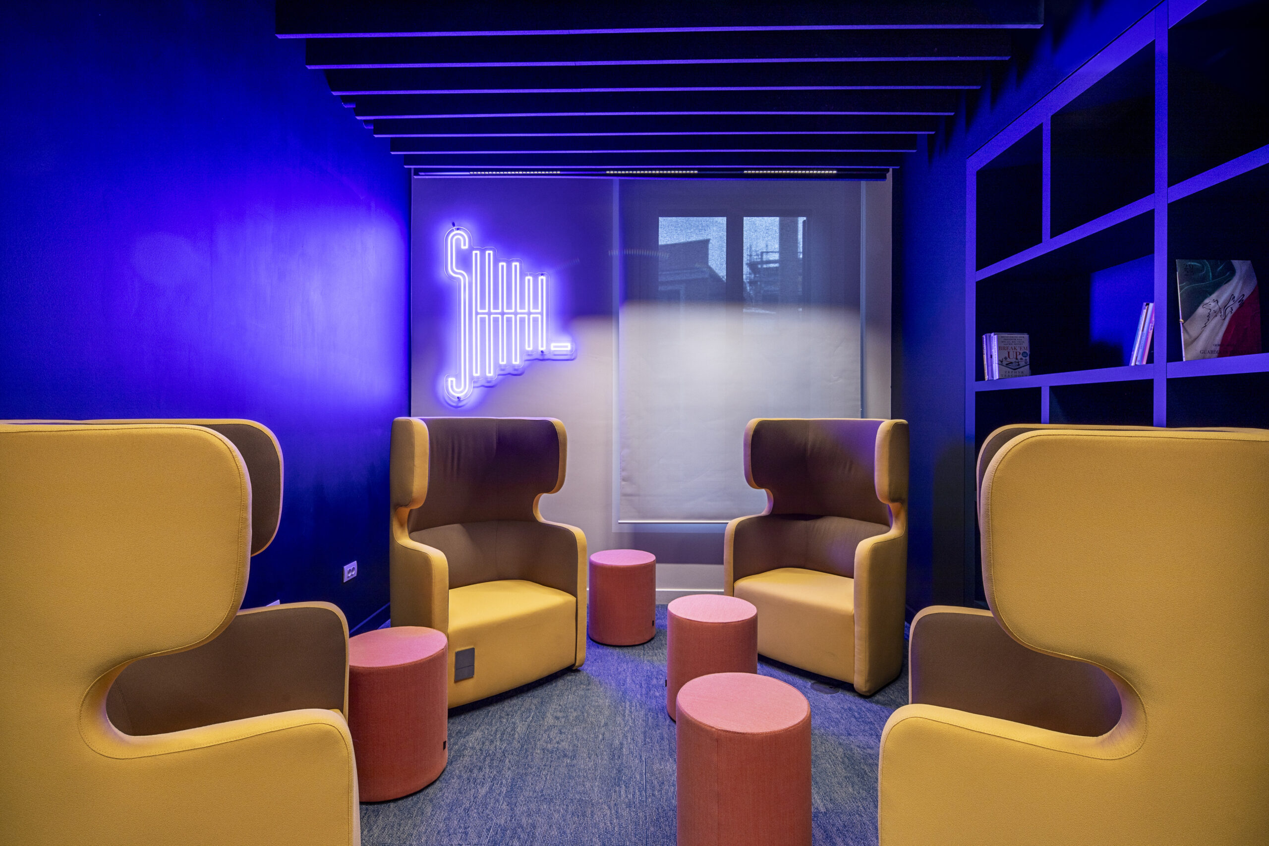Vibrant lounge area featuring bold purple walls and contemporary, sound-absorbing chairs in mustard yellow and brown. Nestled beside a playful neon sign, small round tables in varying heights encourage casual conversation. The design promotes both function and aesthetic appeal, creating an inviting atmosphere for relaxation and engagement.
