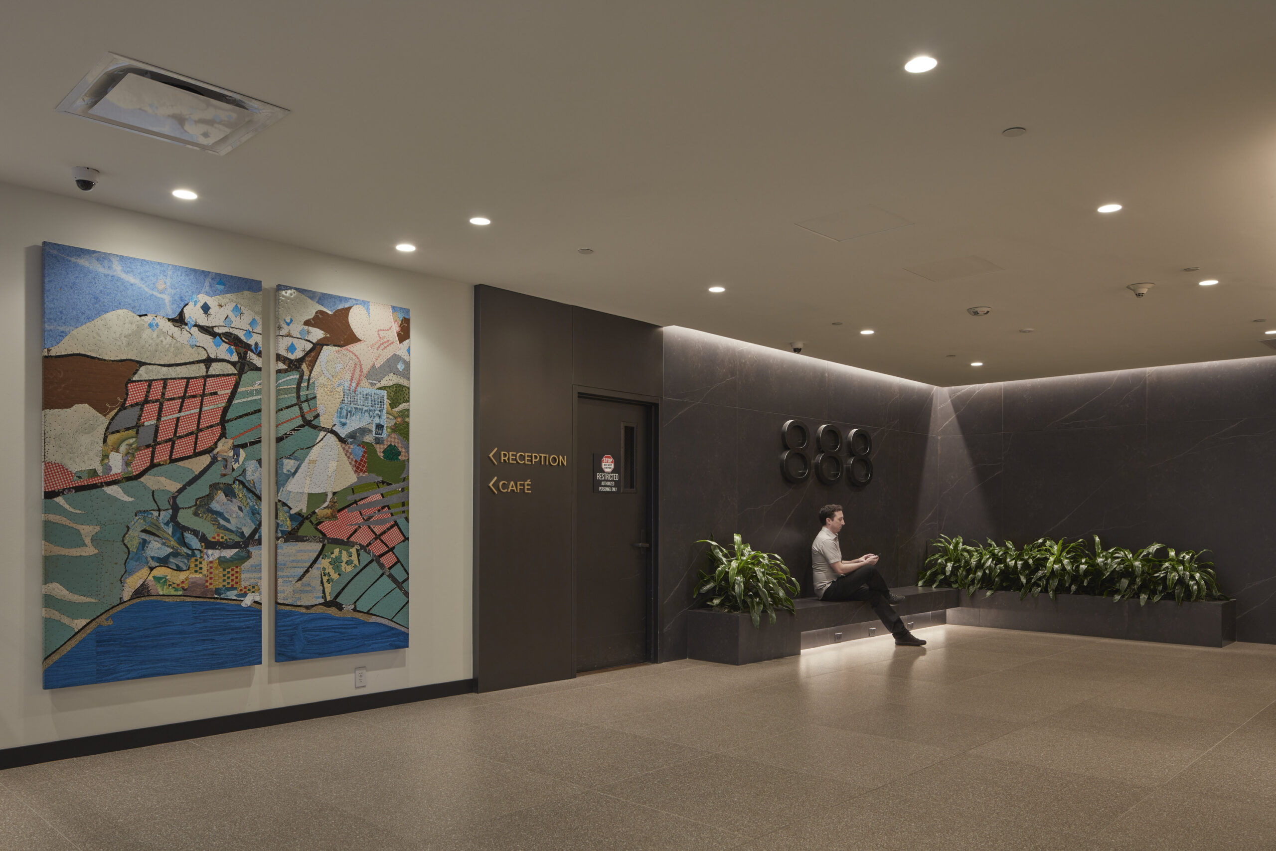 Brightly lit reception area features a large, colorful abstract mural on the left, contrasting with the sleek dark walls. A modern seating nook with integrated greenery provides a cozy spot for relaxation. A man is seated, engaging with his device, embodying the space's inviting atmosphere.