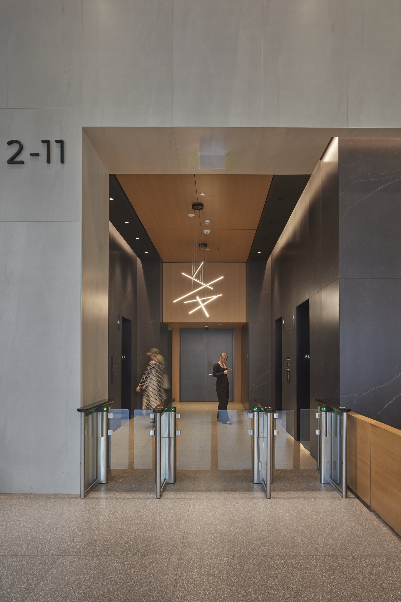 Modern lobby featuring a sleek, minimalistic design with dark stone finishes and warm wooden accents. The space is illuminated by a striking geometric light fixture, enhancing the contemporary aesthetic. Two individuals interact near clear security gates, adding a dynamic element to this bustling entryway.
