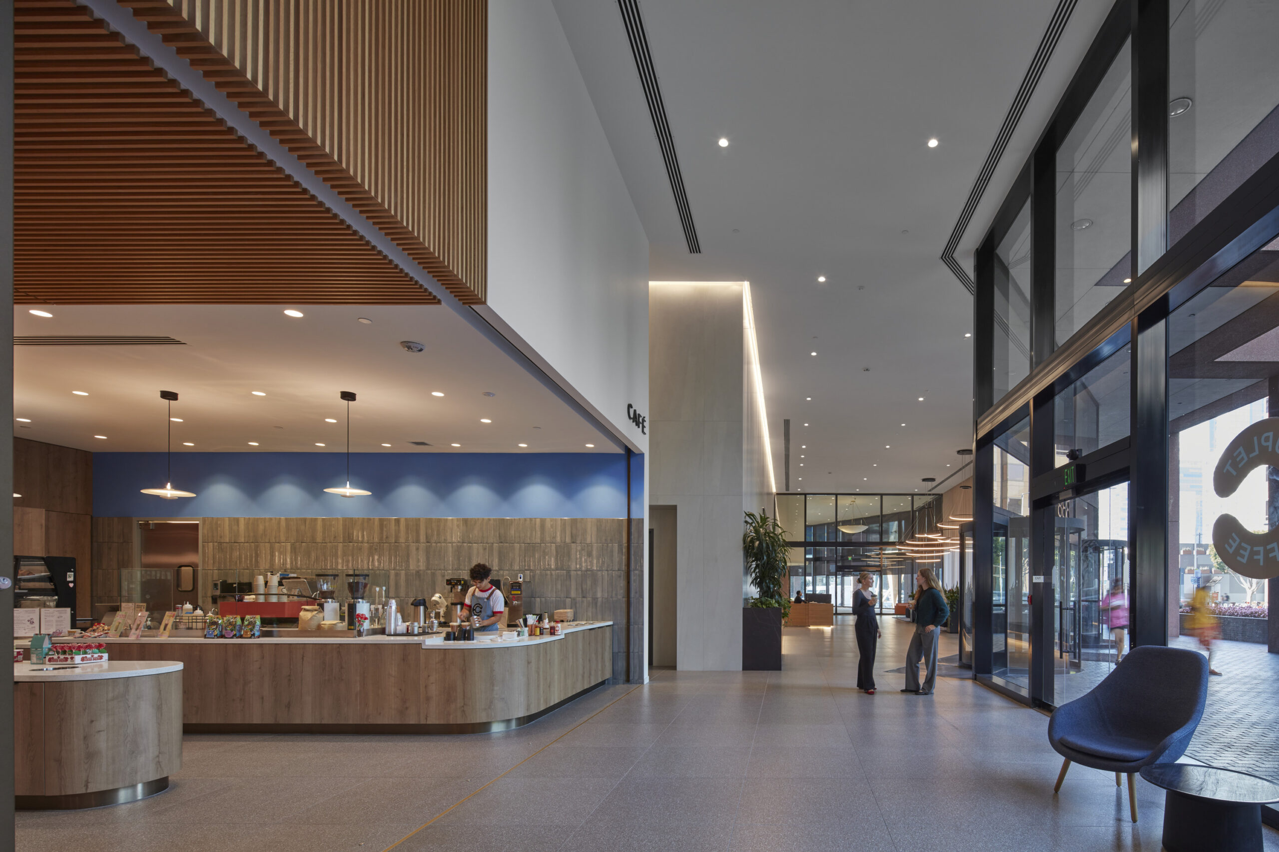 Spacious lobby featuring a modern coffee bar with warm wood accents and blue-gray wall panels. Sleek glass walls allow natural light to enhance the open atmosphere. A sculptural chair adds a touch of elegance, while a subtle ceiling design introduces visual interest to the contemporary space.