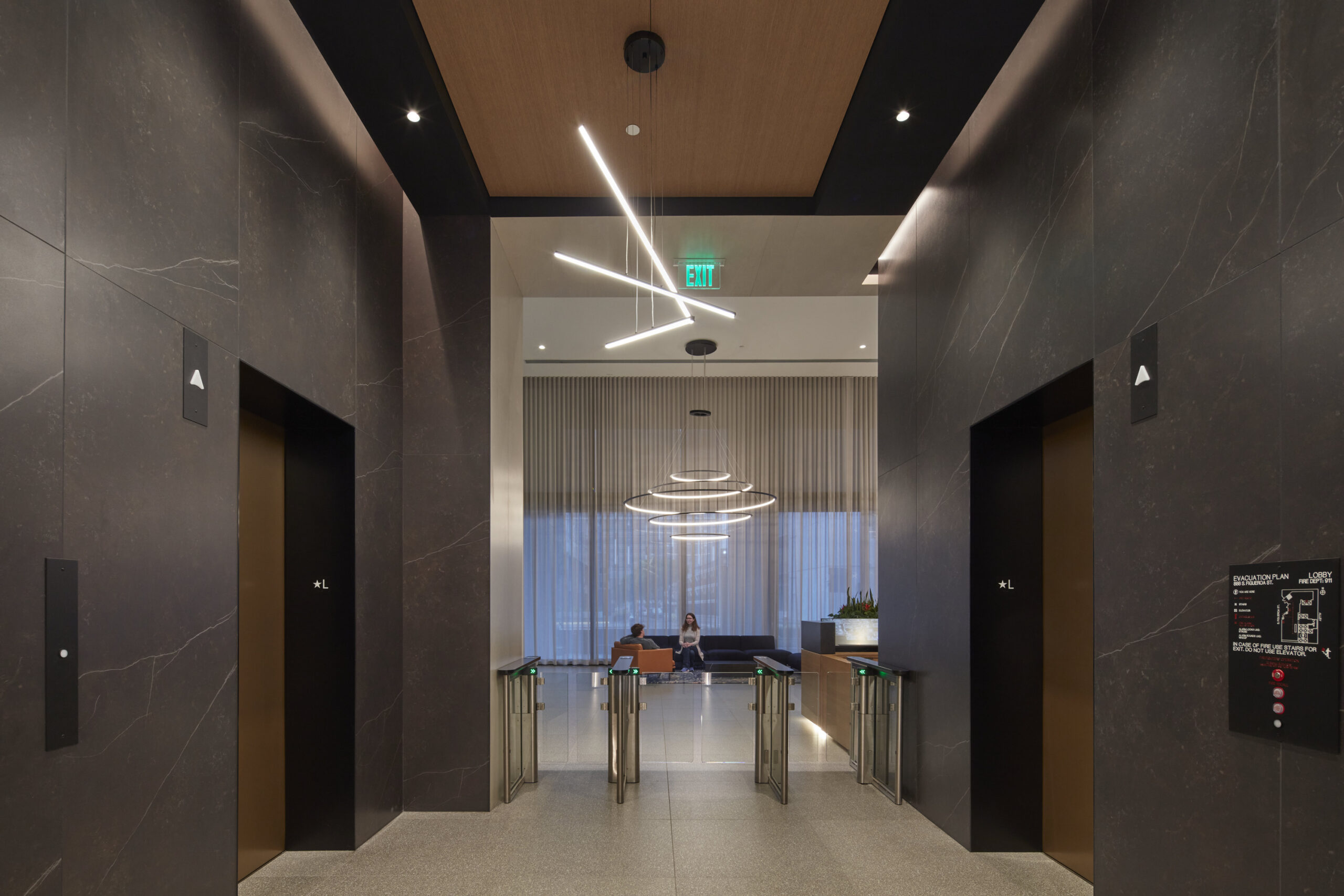 A sleek lobby featuring dark stone walls and a polished concrete floor. Elegant pendant lighting with geometric designs illuminates the space, complementing contemporary elevator doors. Modern furnishings with a neutral palette invite relaxation, while large windows offer sweeping views, enhancing the sophisticated atmosphere of the interior.