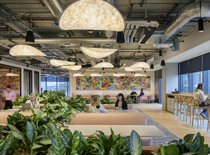 Open office space featuring modern, curved pendant lighting that casts a warm glow. Abundant indoor plants create a lively atmosphere, while a vibrant mural adds a pop of color. Comfortable seating areas encourage collaboration, complemented by sleek, minimalist furniture and large windows allowing natural light to enhance productivity.