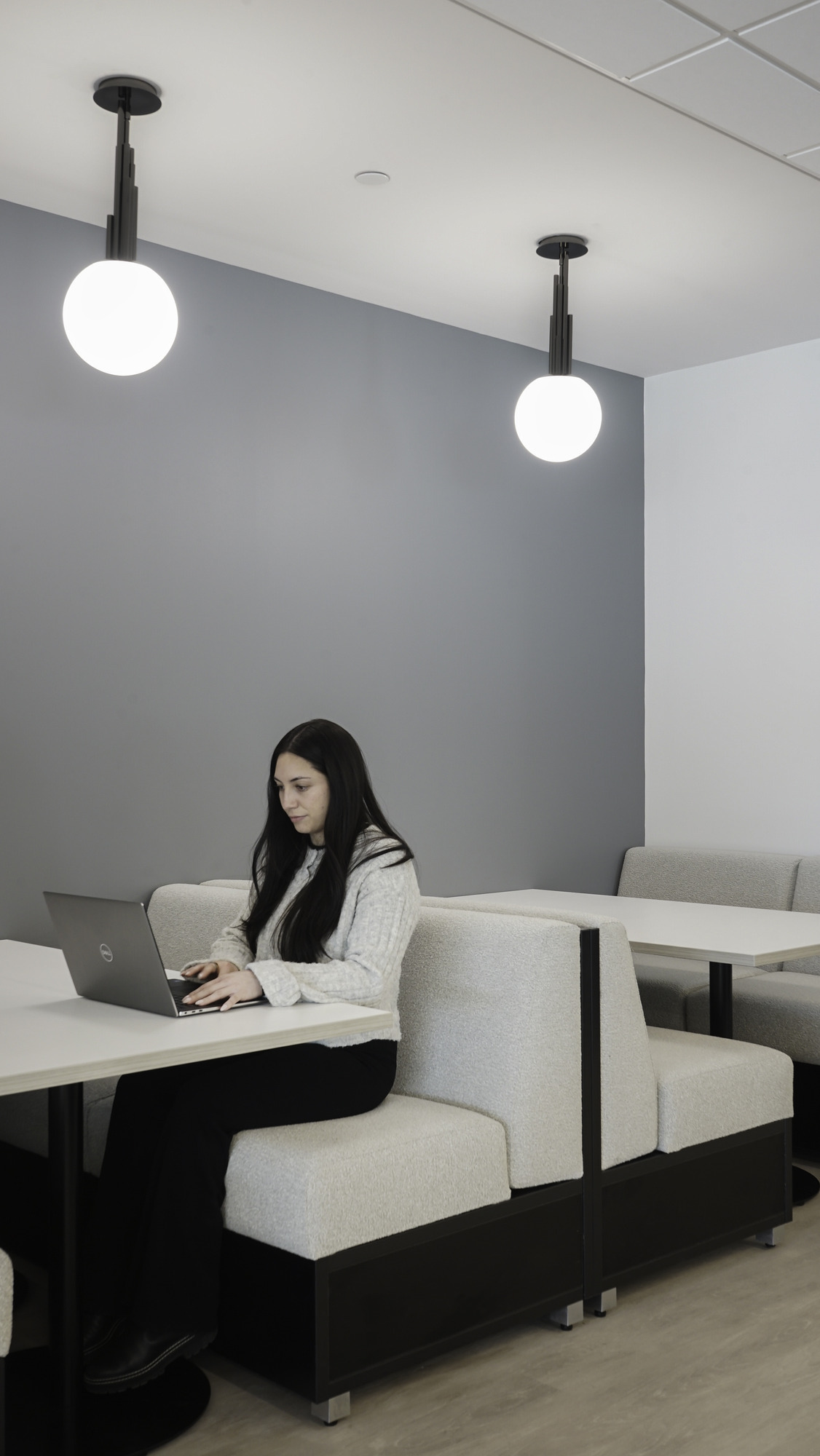 A modern workspace features a woman working on a laptop at a sleek booth seating area. The space is illuminated by two minimalistic pendant lights, contrasting against a soft gray wall. Booths offer a comfortable setting, enhanced by clean lines and a neutral color palette, promoting productivity and collaboration.