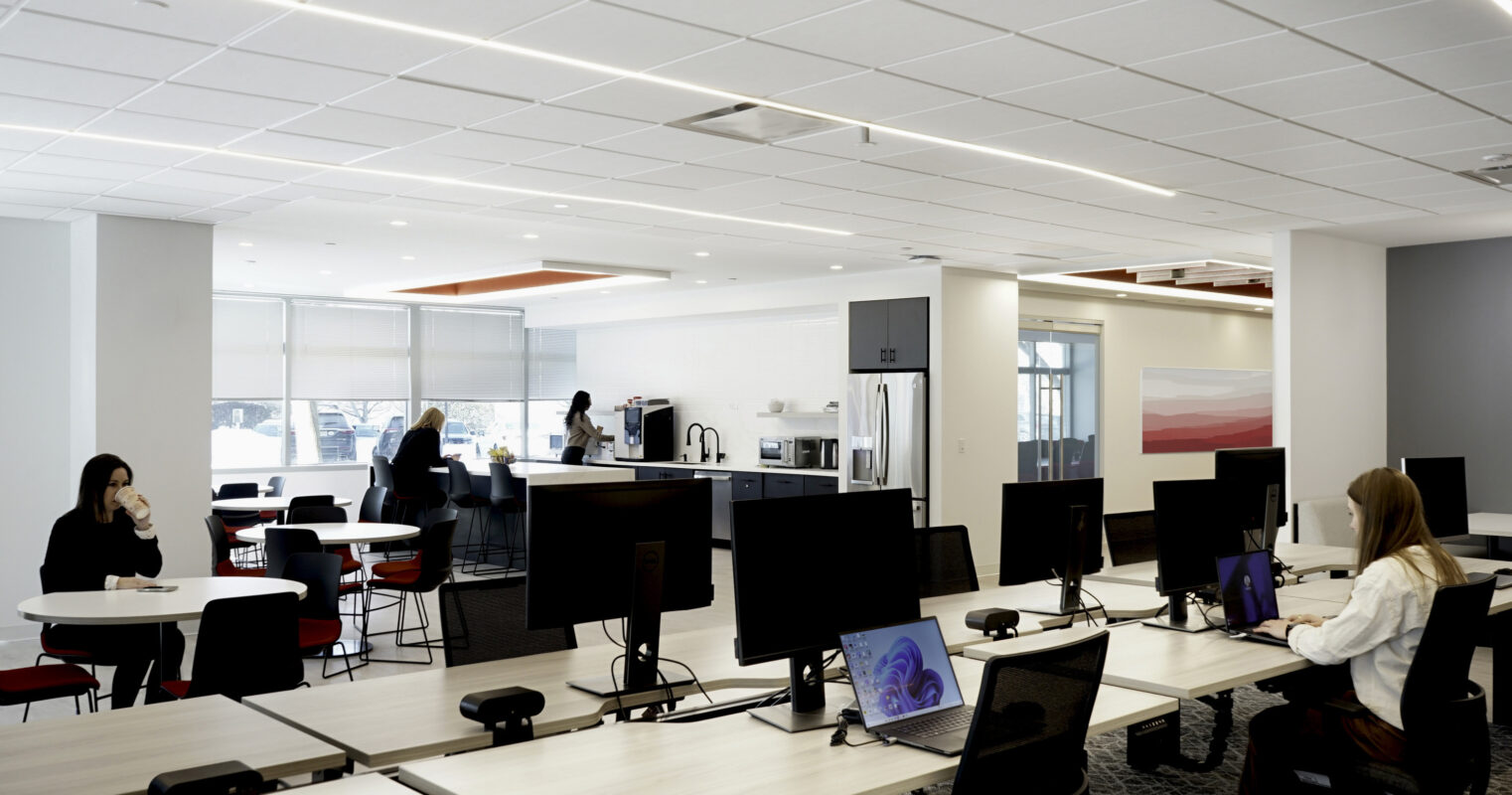 A modern open-plan office features collaborative workspaces with ergonomic desks and sleek computer setups. Natural light floods the area through large windows, complementing minimalist furniture in muted tones. A kitchen area is visible in the background, fostering a balance between productivity and relaxation.