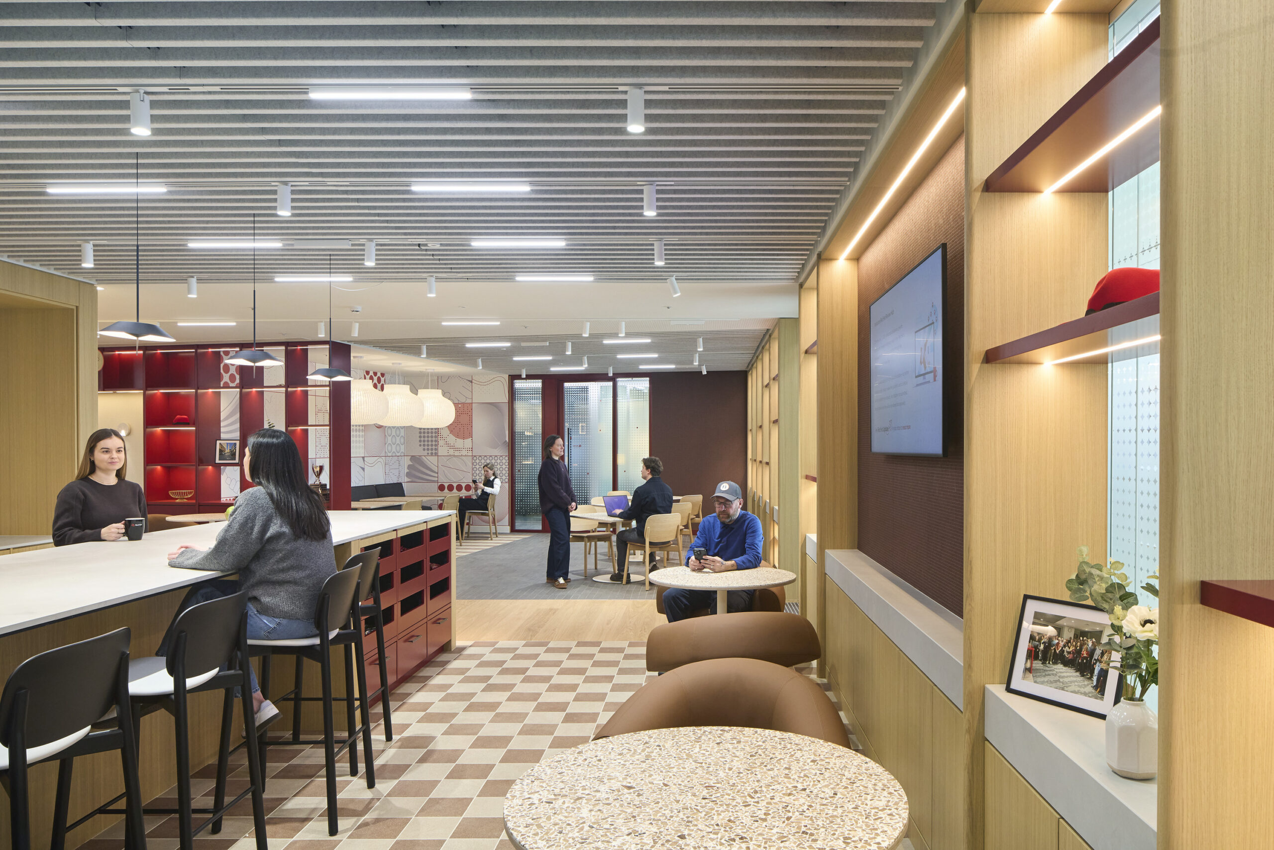 A contemporary office interior features a collaborative workspace with a light-toned wood bar and high-backed stools. Unique design elements include striking patterned wallpaper, whimsical pendant lights, and comfortably arranged seating. The layout encourages both casual interaction and focused work, enhancing a dynamic and inviting professional environment.