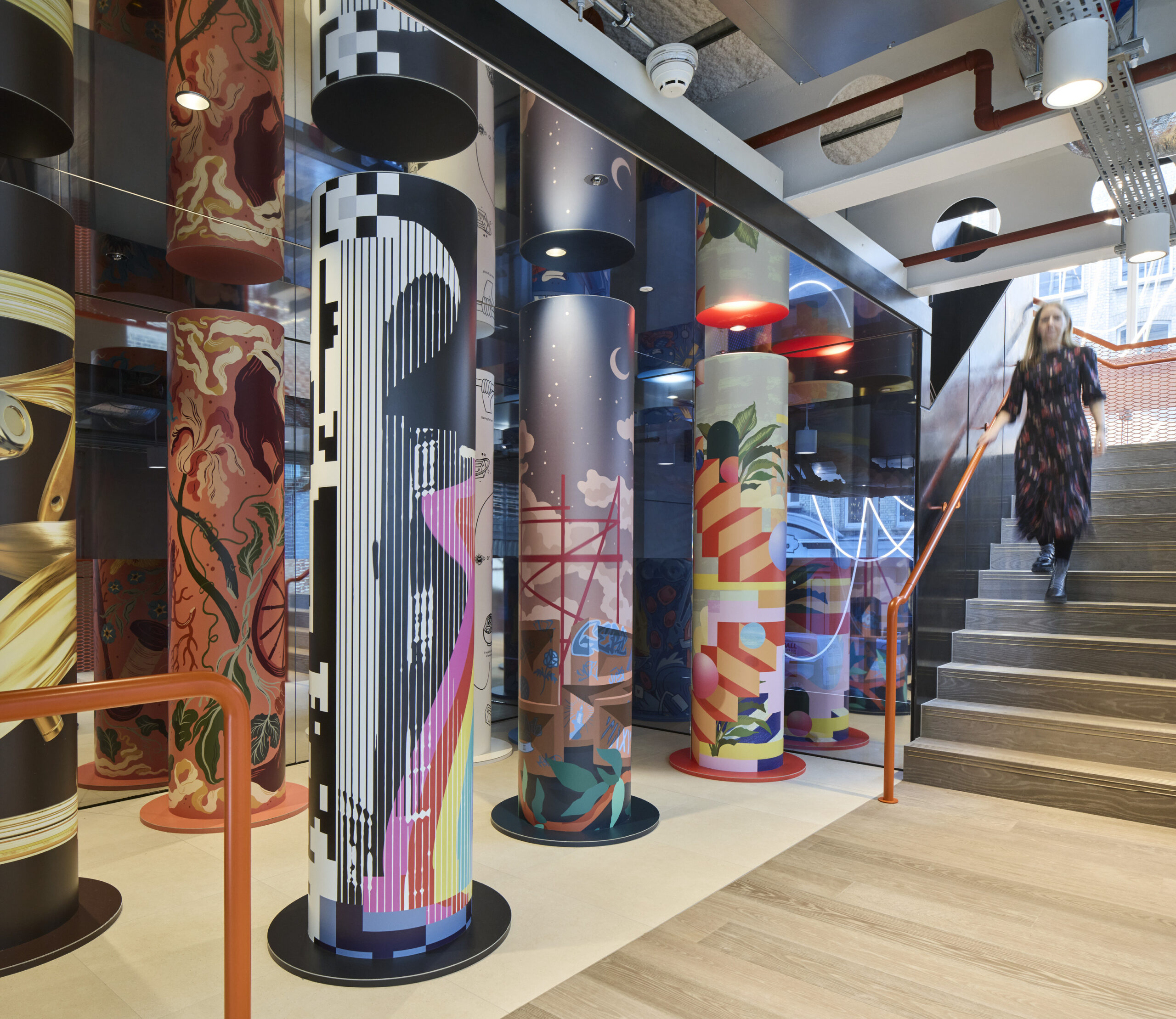 Vibrant, artistically designed columns feature striking graphics and patterns in a modern staircase area, showcasing a blend of contemporary and abstract art styles. The colorful designs enhance the spatial experience, encouraging movement and interaction while integrating visually stimulating elements into the architectural framework. Natural light filters through the space, adding warmth.