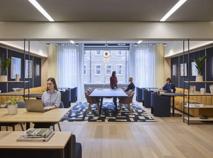 Collaborative office lounge with a central wood meeting table, mustard banquettes, navy lounge chairs, open shelving, framed artwork, plants, and employees working beside a large window.